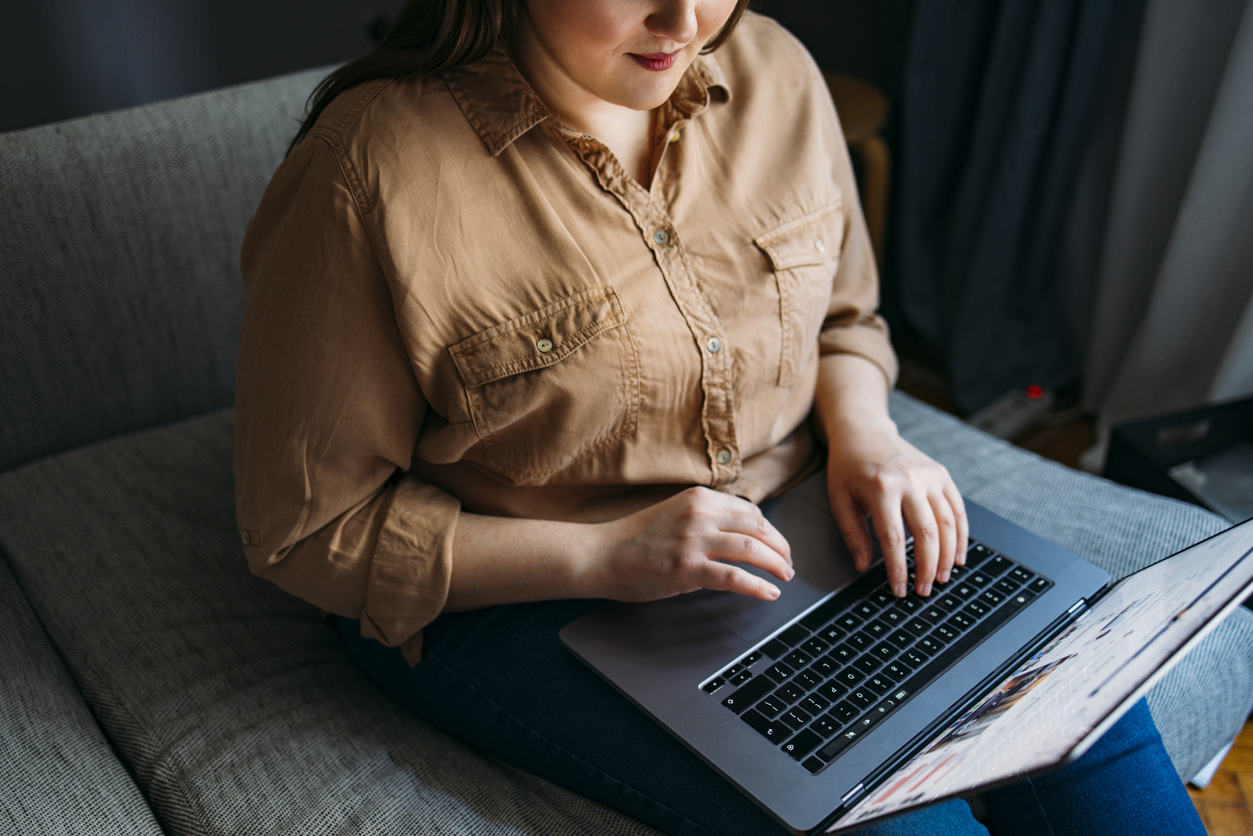 Anonymous Smiling Plus Size Woman Sitting On The Sofa While Working On Her Laptop Unrecognizable Caucasian plus size businesswoman, sitting on a couch while working on her computer