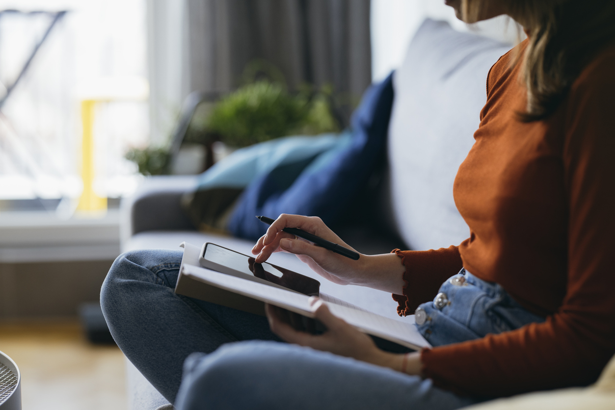 Unrecognizable business woman sitting on couch and using mobile phone while writing notes in notebook in the living room