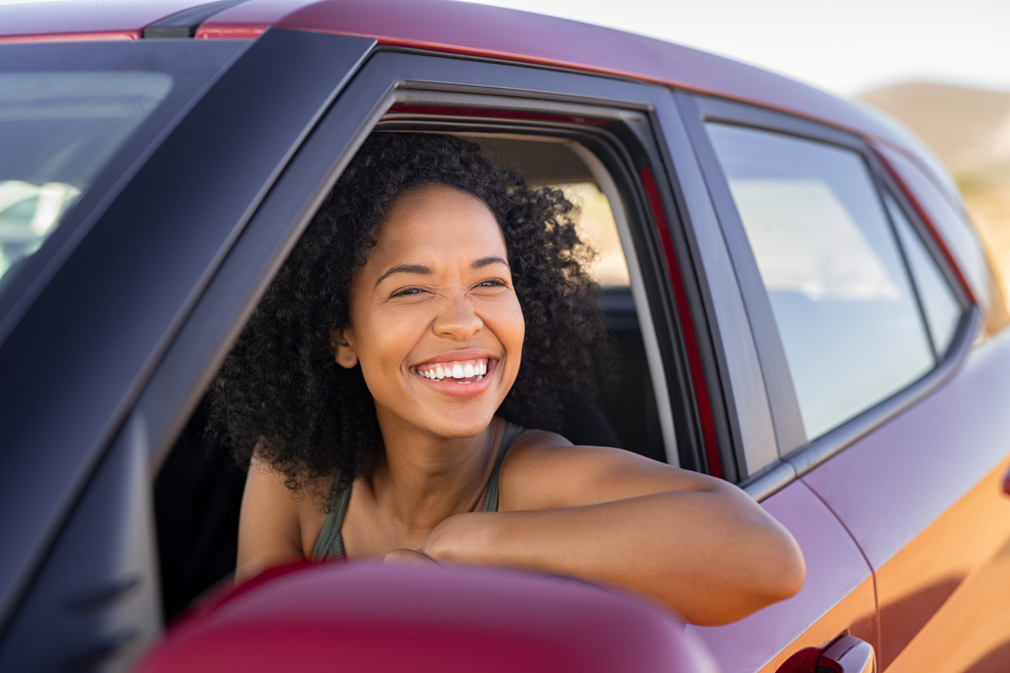 Beautiful african american woman looking outside car during summer holiday. Young attractive black woman laughing and enjoying road trip on a summer day. Happy girl driving a red car with big grin.