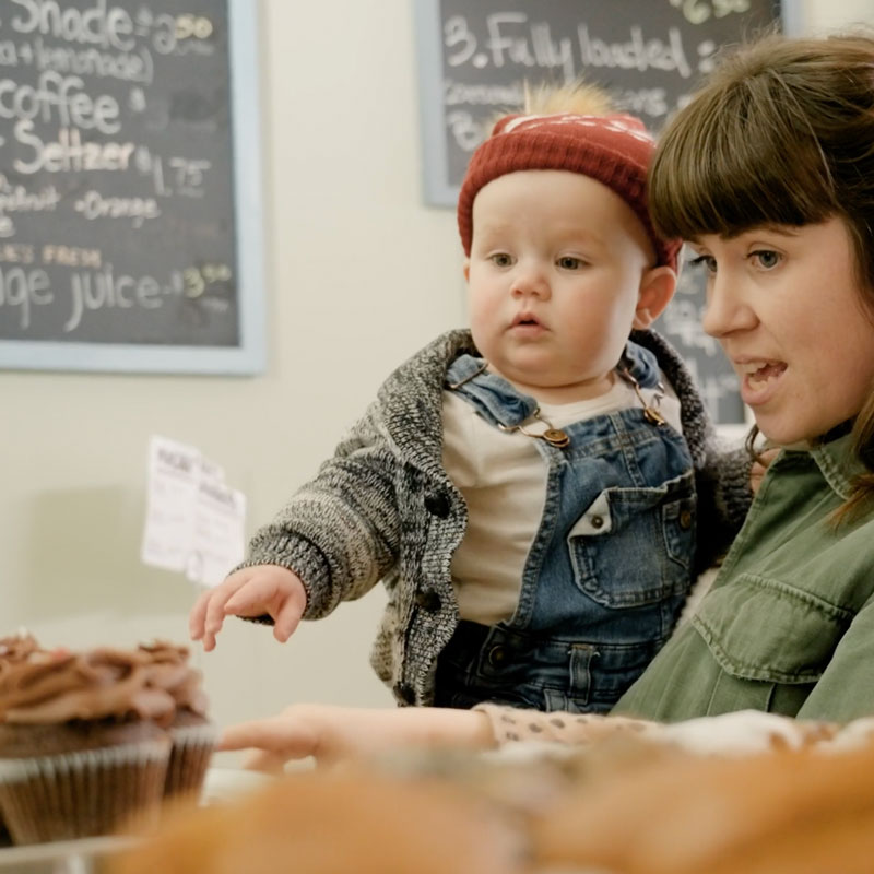 Photo of a woman holding a baby and pointing to treats in at Katie Made Bakery