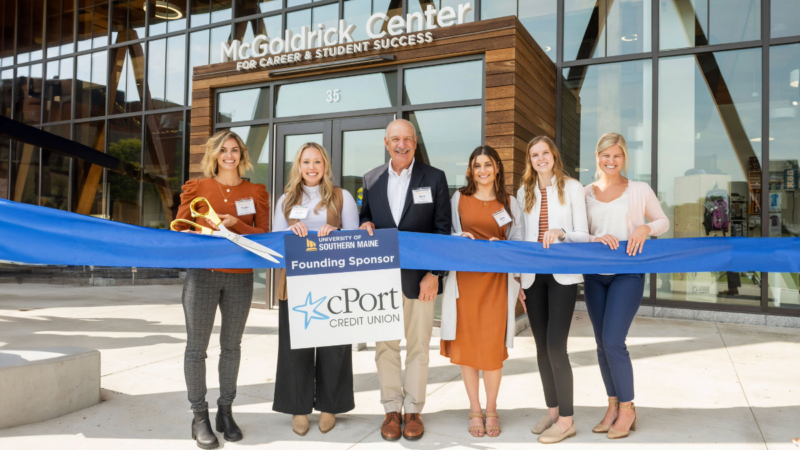 photo of University of Southern Maine alumni and cPort executives standing together in front of USM's McGoldrick Center for Career & Student Success during the ribbon cutting, holding a sign that reads "University of Southern Maine - Founding Sponsor - cPort Credit Union." Pictured in photo are USM Alumni Peyton Collins and Amanda Hill; Gene Ardito, Olivia Haddad, Katherine Morrison, and USM Alum Kelly Chaisson.