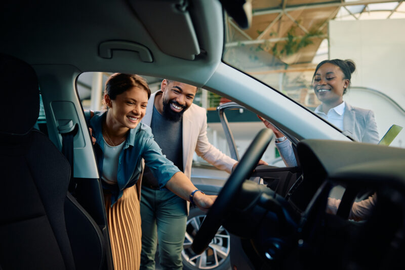 Happy Multiracial Couple Looking At New Car In Showroom.