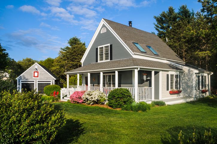 A small Cape Cod style home with a covered wrap around front porch bordered with colorful pink and white azalea bushes on Cape Cod in Massachusetts on a May afternoon. In the background is a detached two car garage