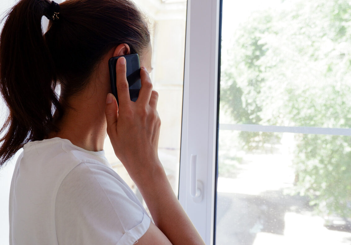 woman with cell phone to her ear while looking out the window of her home