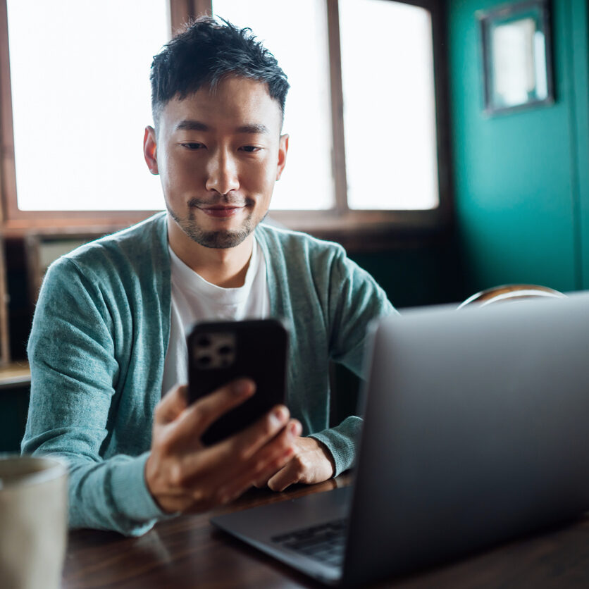 Confident young Asian man looking at smartphone while working on laptop computer in home office. Remote working, freelancer, small business concept
