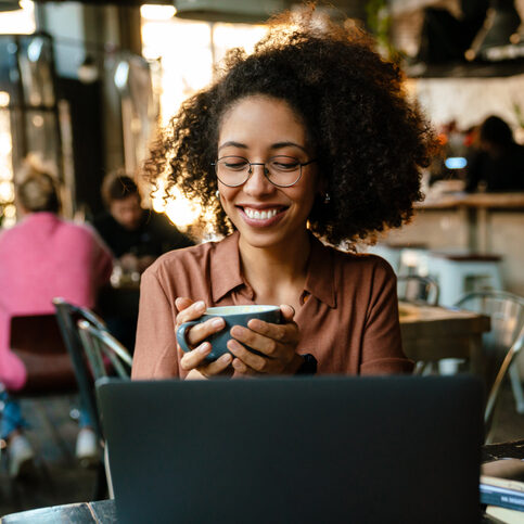 Young african american woman using laptop and drinking coffee while sitting by table in cafe